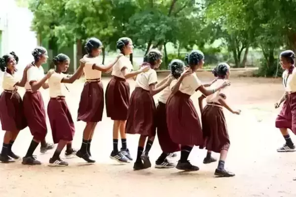 A group of girls wearing matching cream shirts and maroon skirts play together on a dirt schoolyard. They are holding onto each other, leaning back, and laughing. Trees are visible in the background.