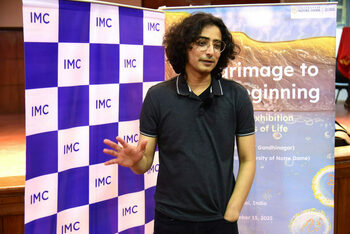 A young man with dark, curly hair and round glasses, wearing a charcoal grey polo, gestures. He stands between a blue and white "IMC" banner and a "University of Notre Dame Global"